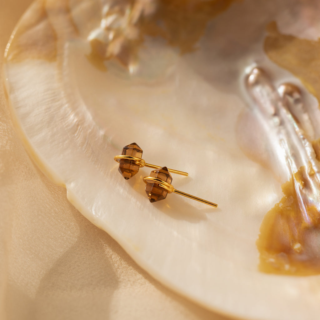 Herkimer Smoky Quartz Studs—gold stud earrings featuring brown gemstones—are displayed on a pearlescent shell under soft, warm lighting.
