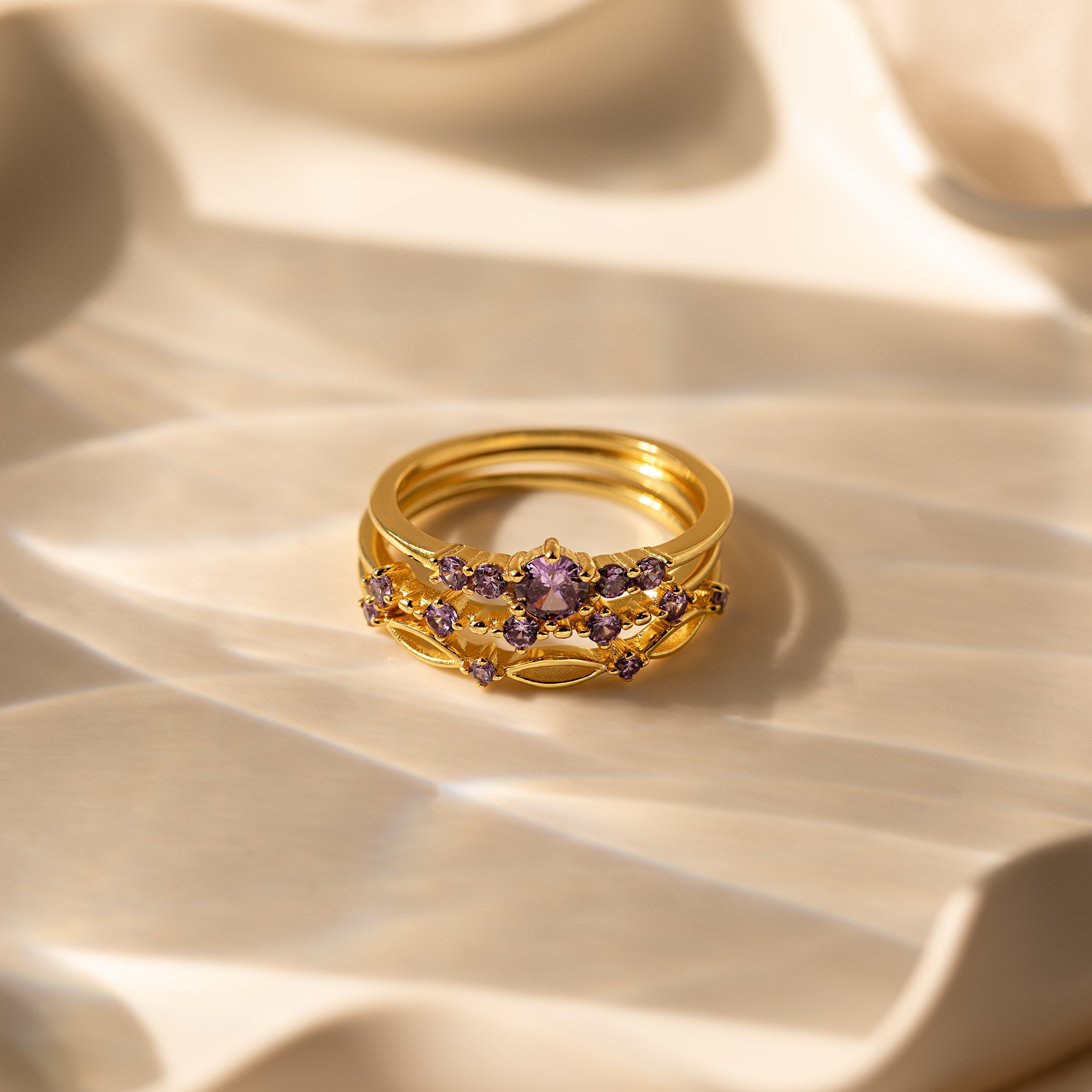 Gold rings, including the Amethyst Stacking Ring Set, are displayed on a softly textured cream surface under gentle light and shadows.