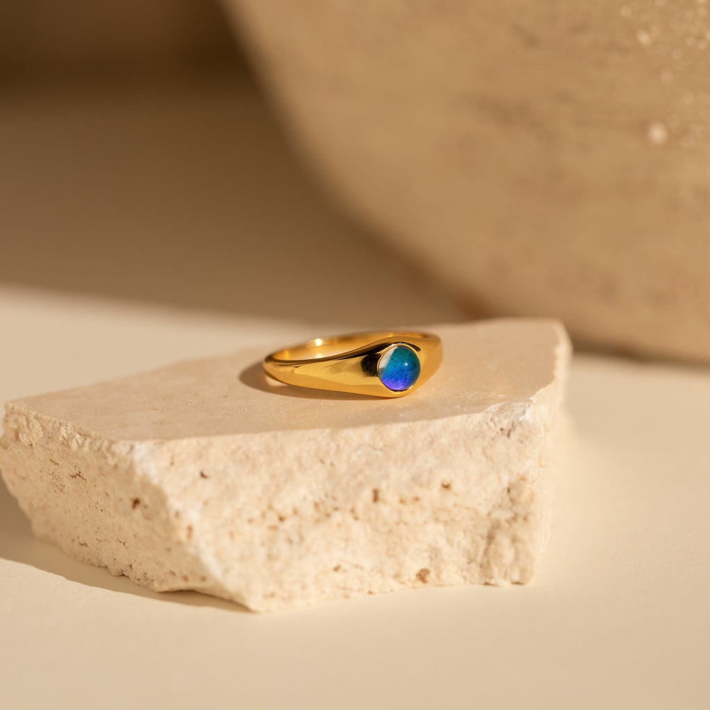 The Mini Signet Mood Ring, featuring a minimalist design and a blue stone, sits on light-colored stone in gentle natural light—ideal for stacking or wearing alone for a subtle look.
