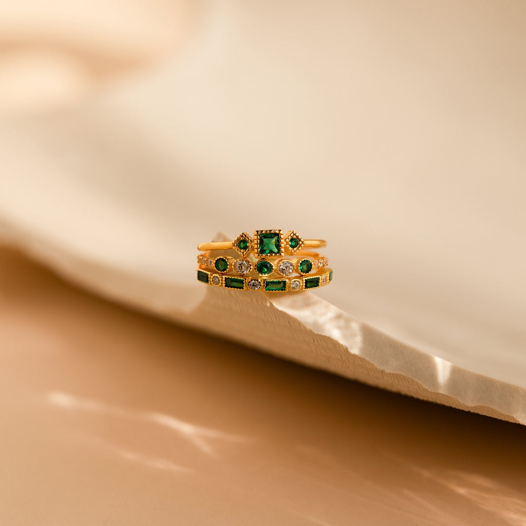 The Emerald Stacking Ring Set in 18K Gold features vintage-inspired green and clear gemstones, evoking art deco charm and elegance, beautifully displayed on a textured, light-colored surface.