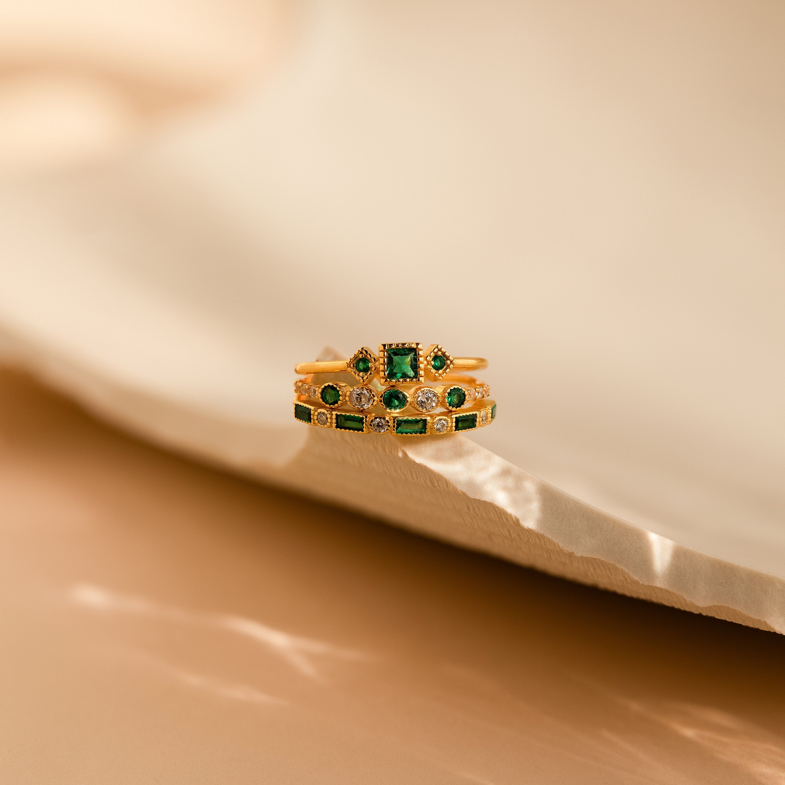 The Emerald Stacking Ring Set in 18K Gold features vintage-inspired green and clear gemstones, evoking art deco charm and elegance, beautifully displayed on a textured, light-colored surface.