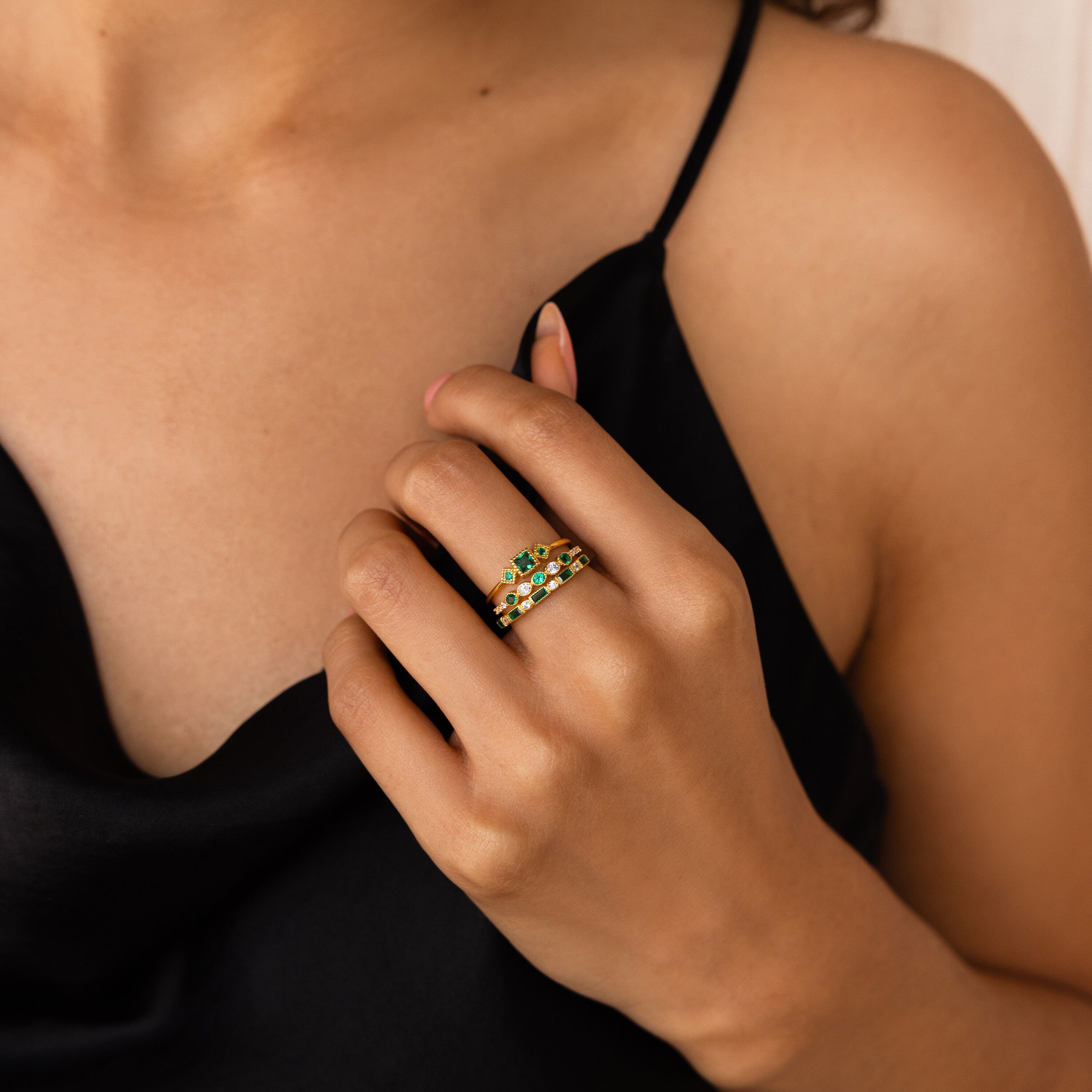 A woman in a black dress touches her chest, wearing gold rings with green gemstones, including the Emerald Stacking Ring Set that adds vintage, art deco-inspired elegance to her look.