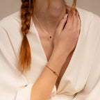 A woman with a braid wears a white robe, a gold necklace with a green gem pendant, and the Emerald Baguette Tennis Bracelet to add statement flair to her elegant look.