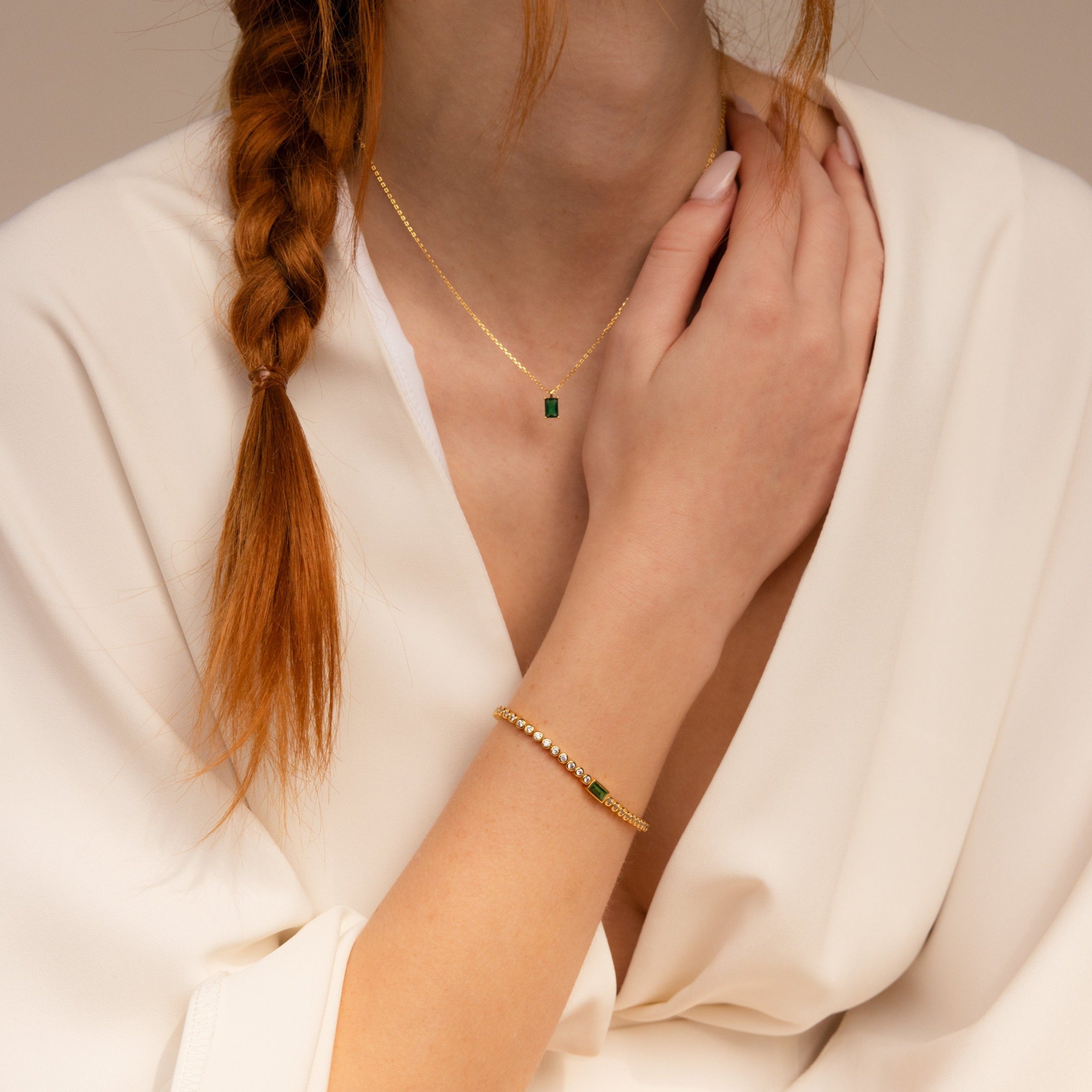 A woman with a braid wears a white robe, a gold necklace with a green gem pendant, and the Emerald Baguette Tennis Bracelet to add statement flair to her elegant look.