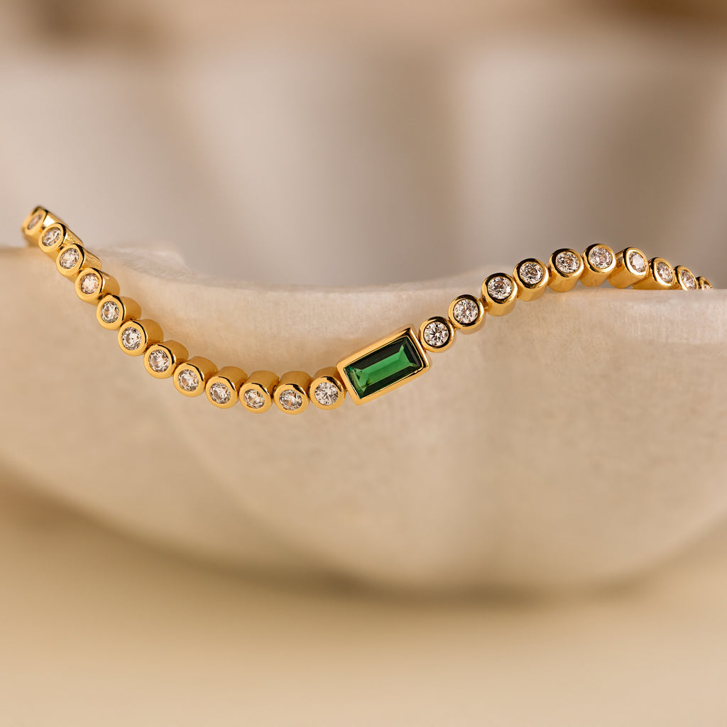 The Emerald Baguette Tennis Bracelet showcases round CZ Diamond gemstones and a green baguette-cut stone, styled on a soft beige background—a perfect statement piece for any event.