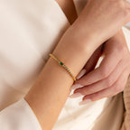 A woman's hand showcases the Emerald Baguette Tennis Bracelet, featuring green gemstones and CZ diamonds, paired with a cream outfit.