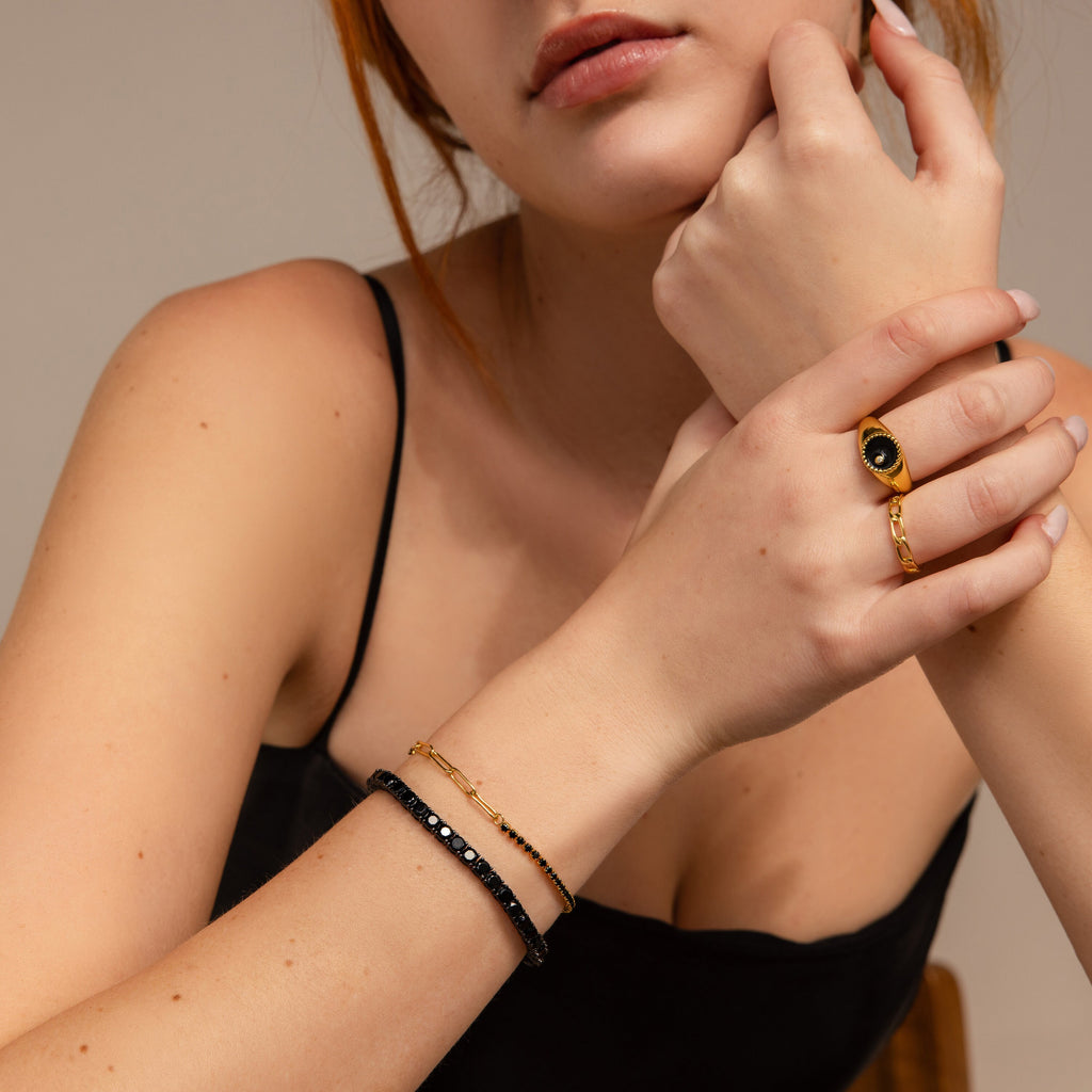 A woman in a black top poses with her hand on her chin, showcasing a gold ring with a black stone and the Onyx Tennis Bracelet.