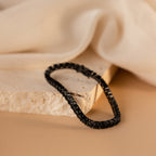 The Onyx Tennis Bracelet, featuring black CZ diamonds, is displayed on a textured stone surface with a soft beige fabric background.