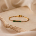 The Emerald Diamond Tennis Bracelet features vibrant emerald gemstones and sparkling diamonds, showcased on a textured stone surface with a soft fabric backdrop.