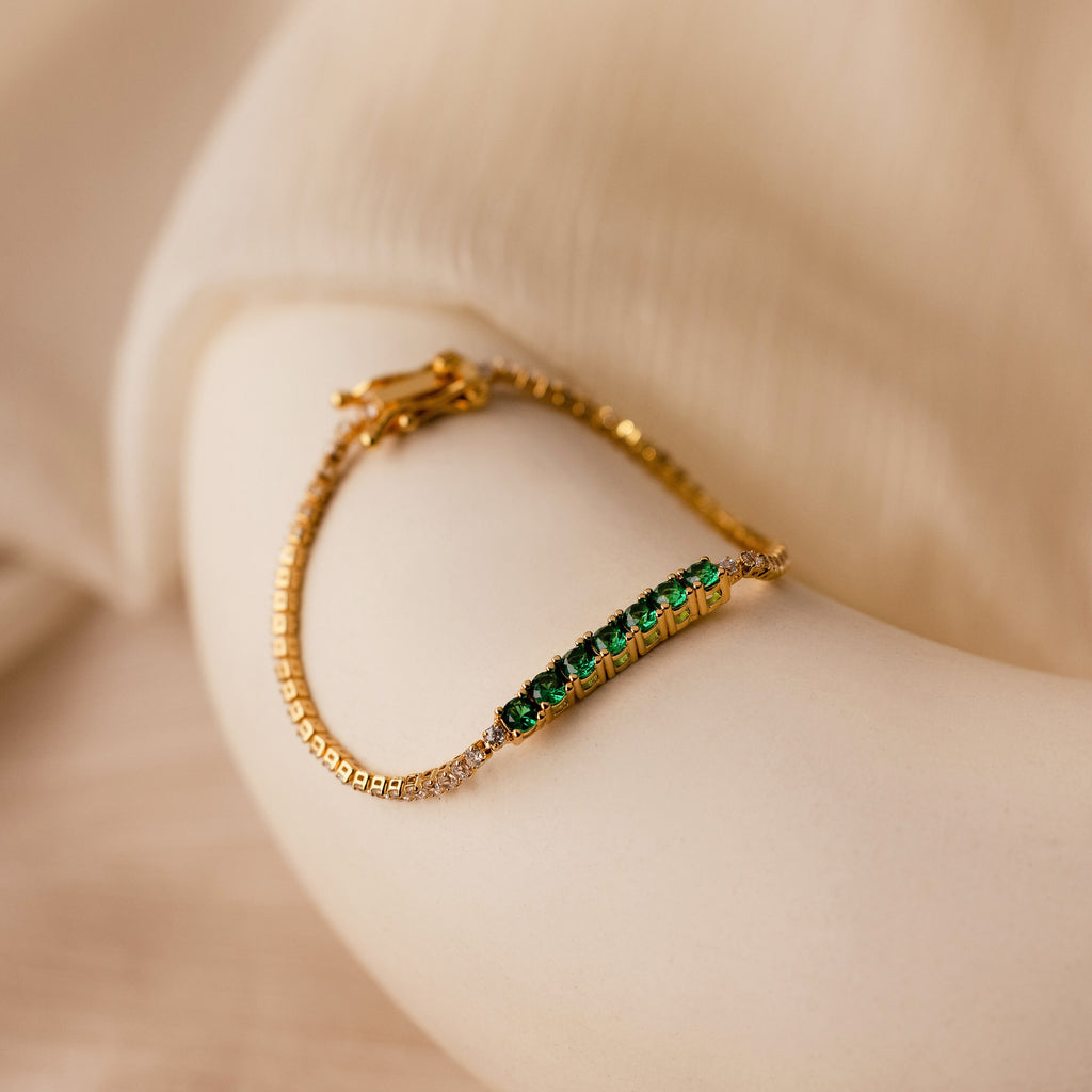 The Emerald Diamond Tennis Bracelet features green emerald gemstones and clear diamonds, elegantly displayed on a smooth, cream-colored curved surface.