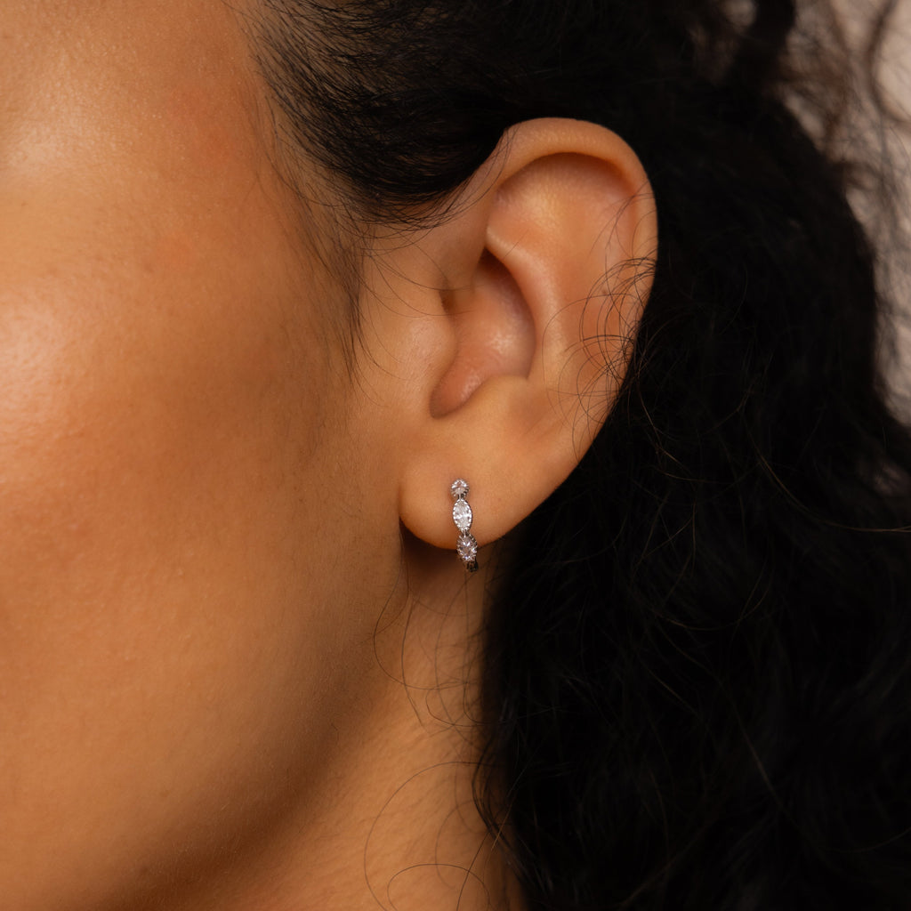A close-up of a woman's ear wearing Marquise Inlay Huggies—small silver hoop earrings, each adorned with three sparkling stones for an elegant touch.