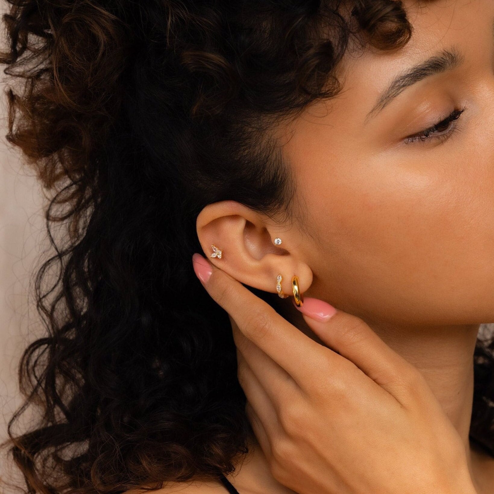A woman with curly hair touches her ear, wearing multiple gold and diamond earrings, including glimmering huggie hoops and Marquise Inlay Huggies.