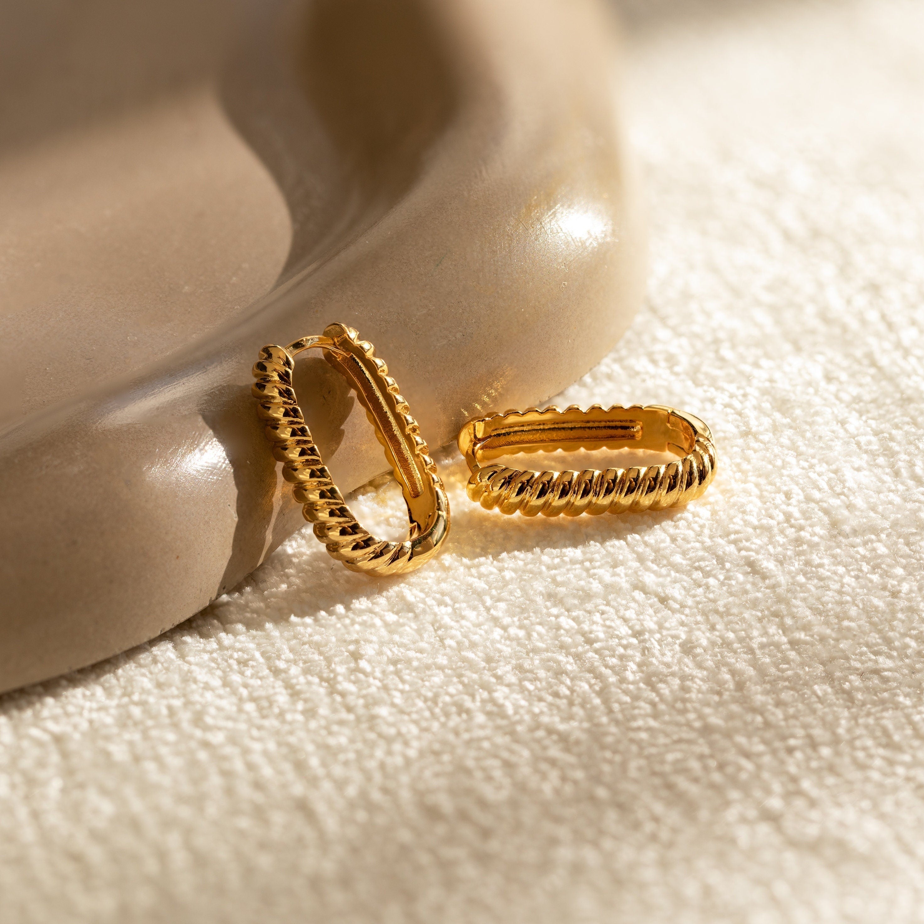 Two Twisted Link Hoops featuring a trendy, ribbed, twisted design are displayed on a soft fabric-textured tan surface.