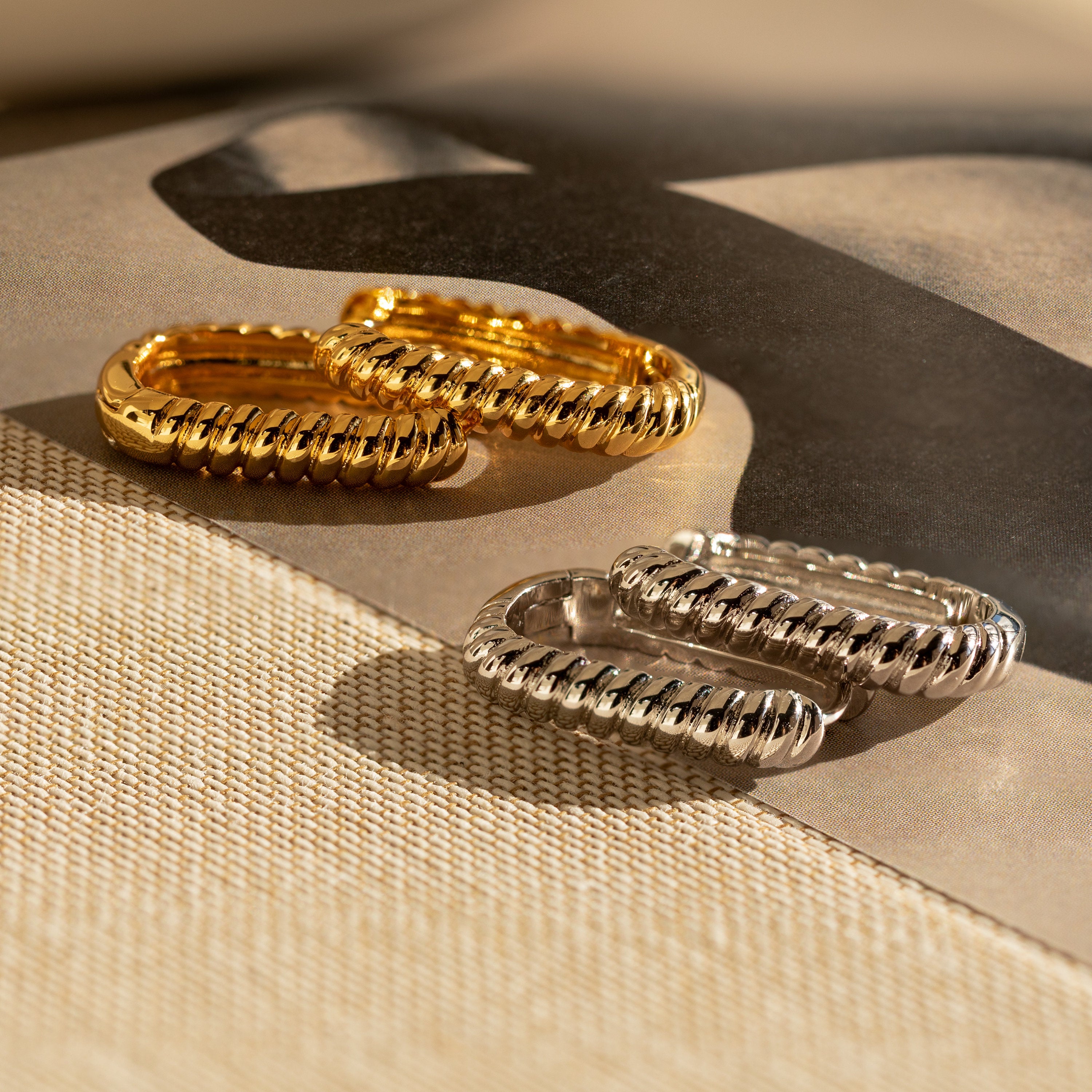 Two pairs of Twisted Link Hoops—one gold, one silver—with textured oval designs are displayed on a neutral, textured surface.