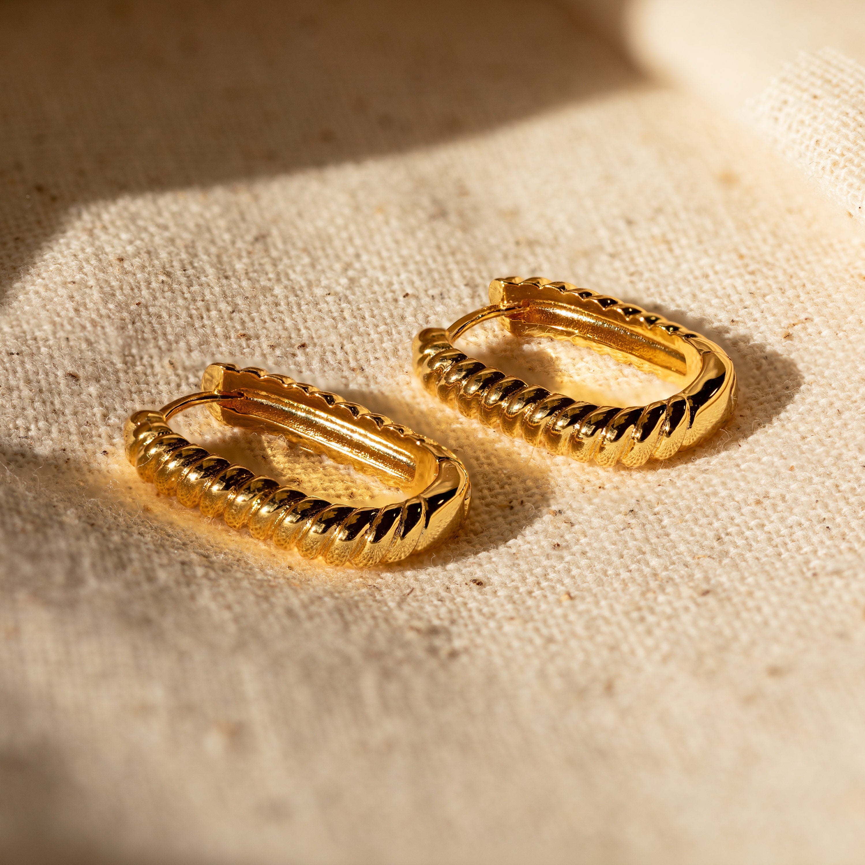 Twisted Link Hoops displayed on beige fabric in warm sunlight—these trendy earrings bring modern elegance to any look.