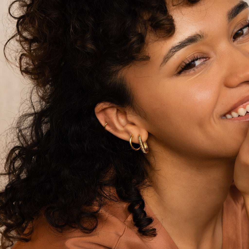 Smiling woman with curly hair wears Twisted Link Hoops and a brown top, resting her hand on her chin.