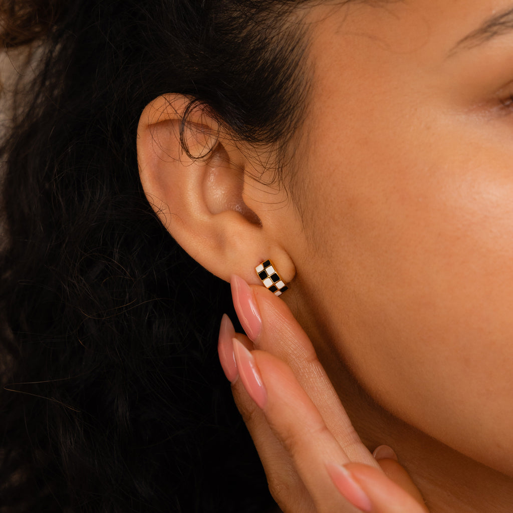 Close-up of a person’s ear wearing Checkered Huggies, stylish black and white earrings, with a hand gently touching the earlobe.