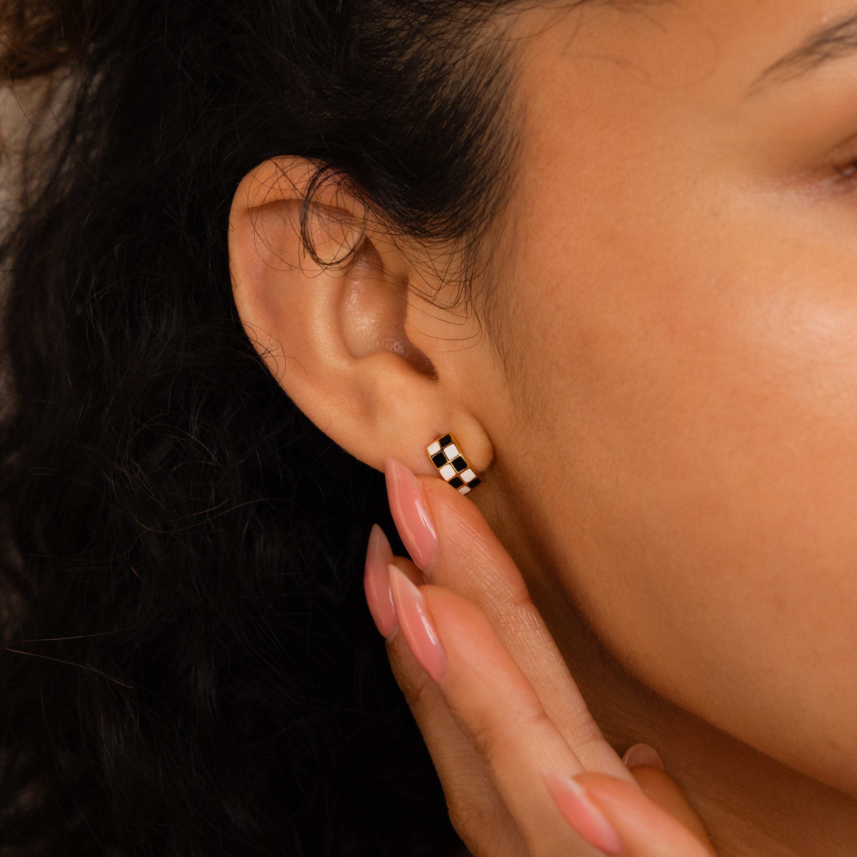 Close-up of a person’s ear wearing Checkered Huggies, stylish black and white earrings, with a hand gently touching the earlobe.