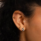 A close-up of an ear wearing four earrings: a black stud, pearl stud, gold hoop, and Checkered Huggies—black and white hoops that bring a modern edge.