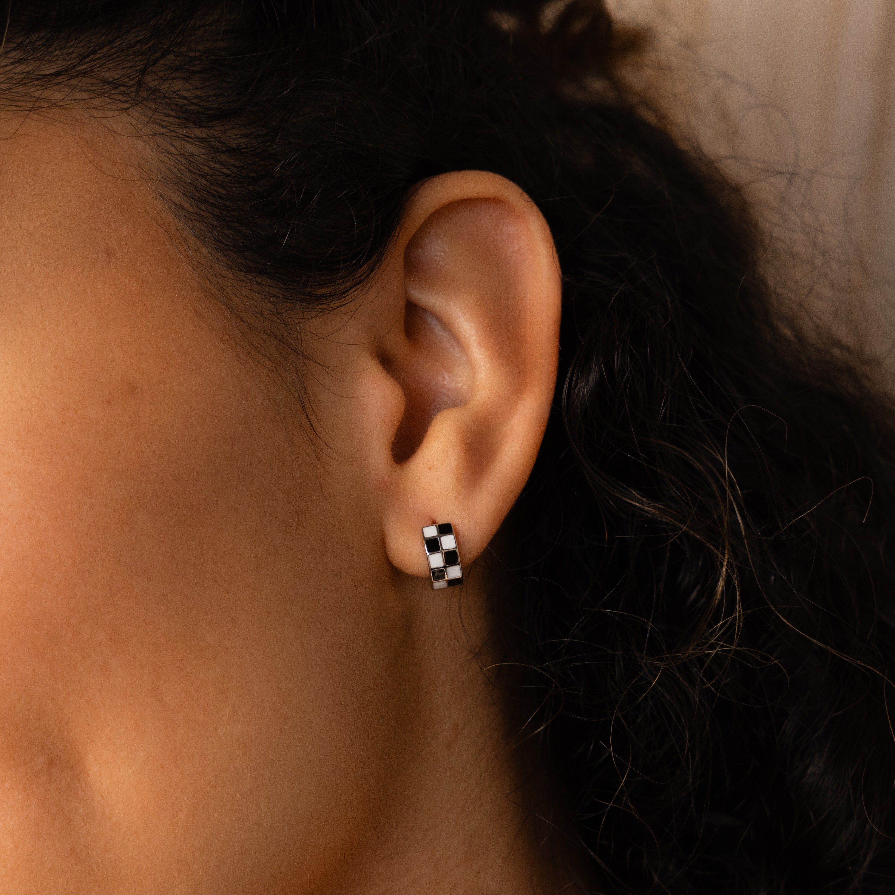A close-up of an ear wearing Checkered Huggies—black and white hoop earrings with a trendy checkerboard design.
