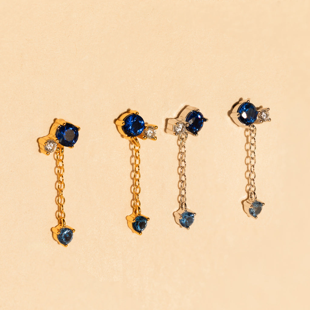 The Sapphire Diamond Drop Earrings feature blue and clear stones set in gold and silver, displayed on a beige background.