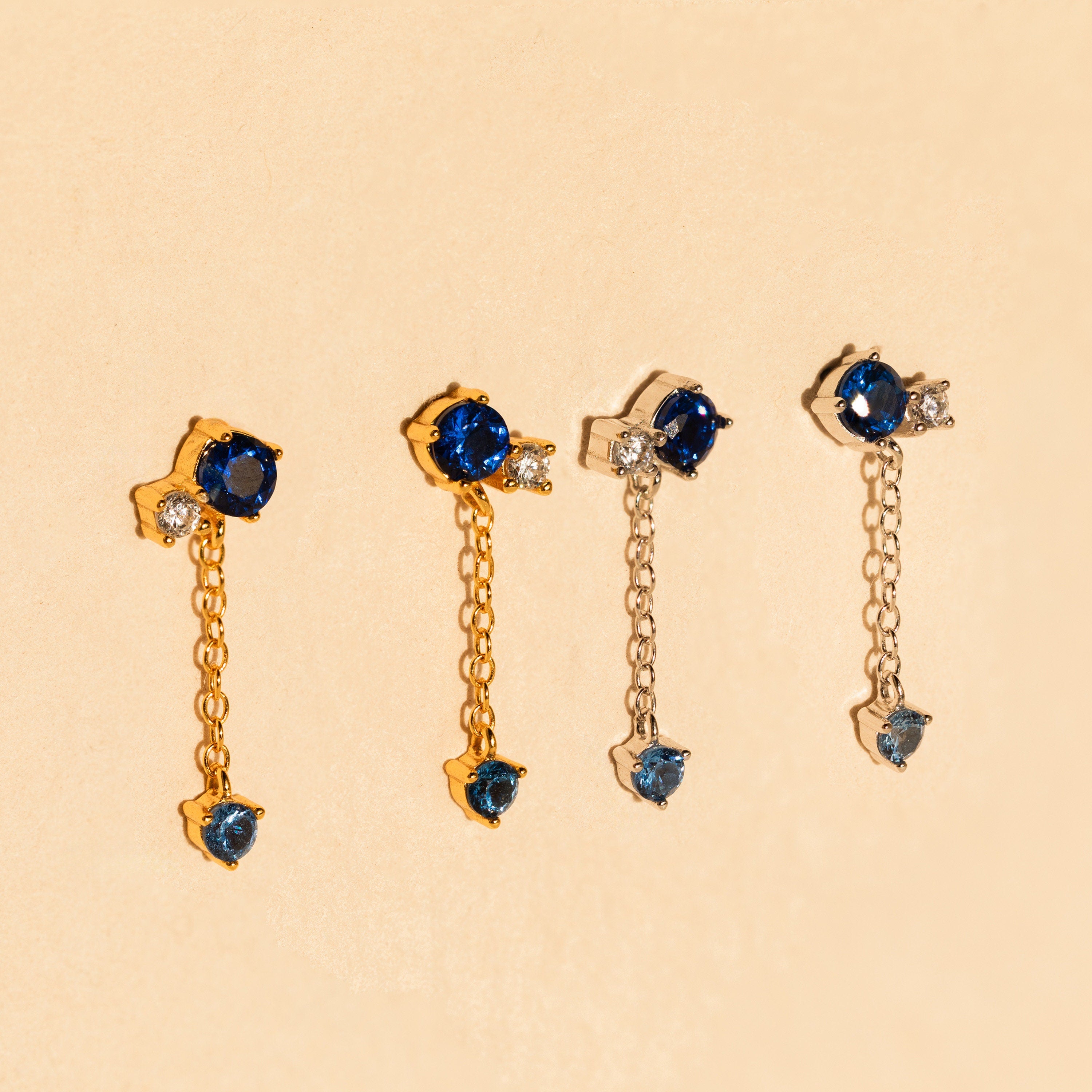 The Sapphire Diamond Drop Earrings feature blue and clear stones set in gold and silver, displayed on a beige background.