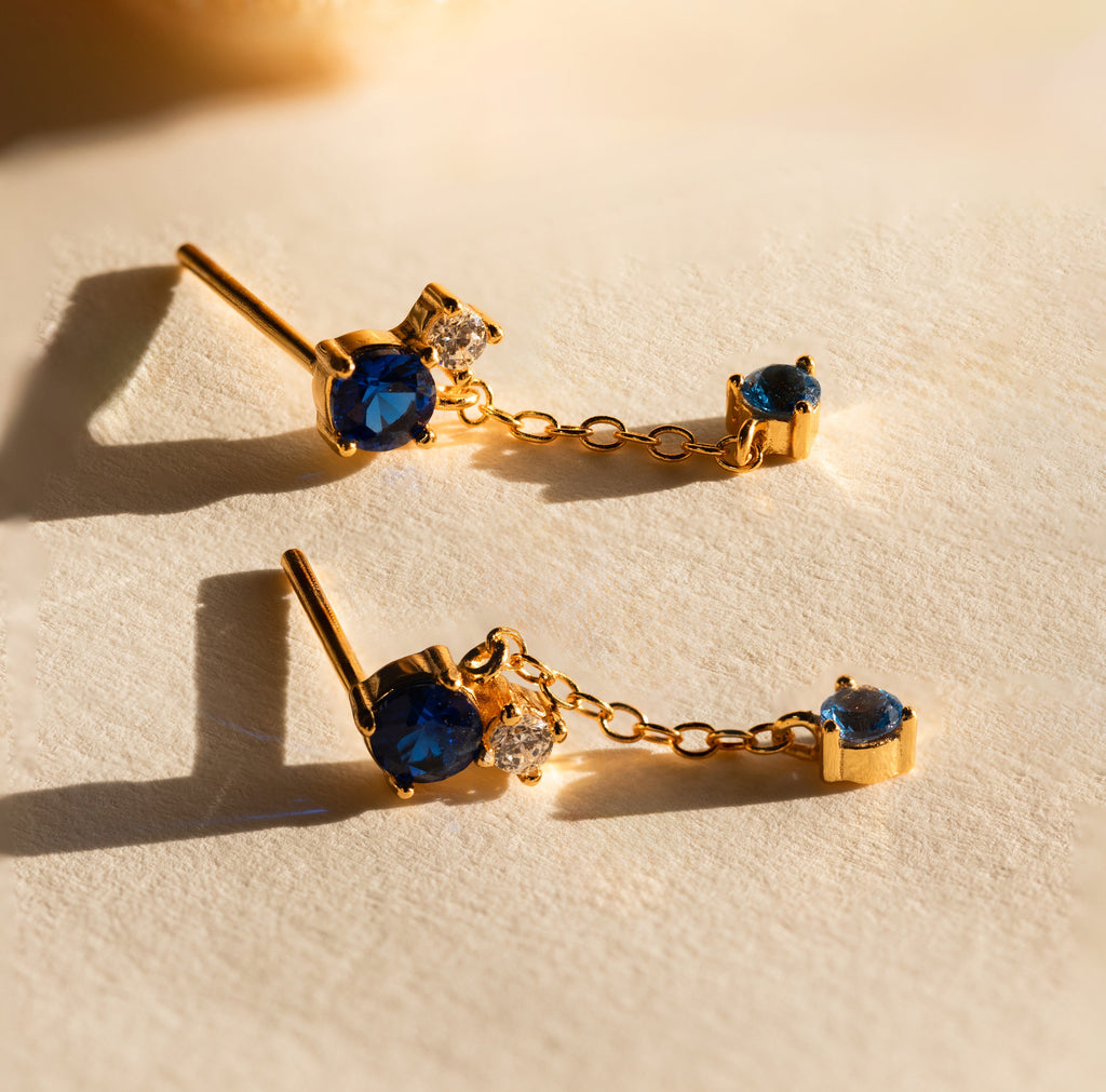 The Sapphire Diamond Drop Earrings feature gold with blue sapphires and clear diamonds, connected by a delicate chain and displayed on a light textured surface.