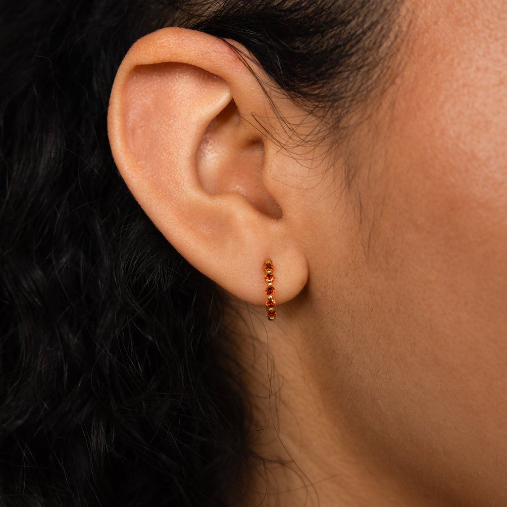 A close-up of an ear wearing the Inside Out Huggies Set, featuring small gold gemstone hoops with red stones, highlighting the elegance of birthstone earrings.