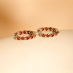 The Inside Out Garnet Huggies, featuring vivid garnet stones set in silver hoops, are displayed on a smooth beige surface against a soft background.