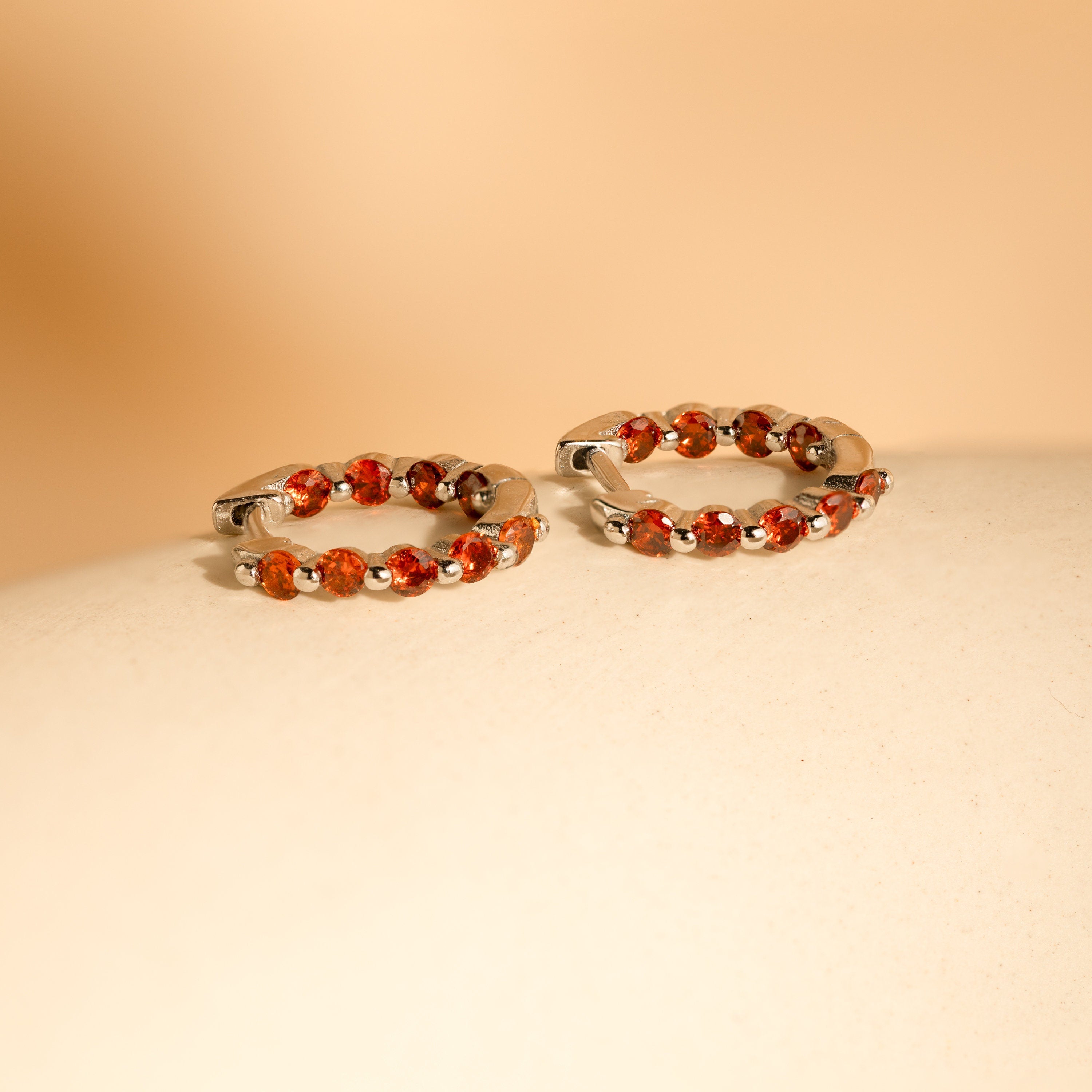 The Inside Out Garnet Huggies, featuring vivid garnet stones set in silver hoops, are displayed on a smooth beige surface against a soft background.