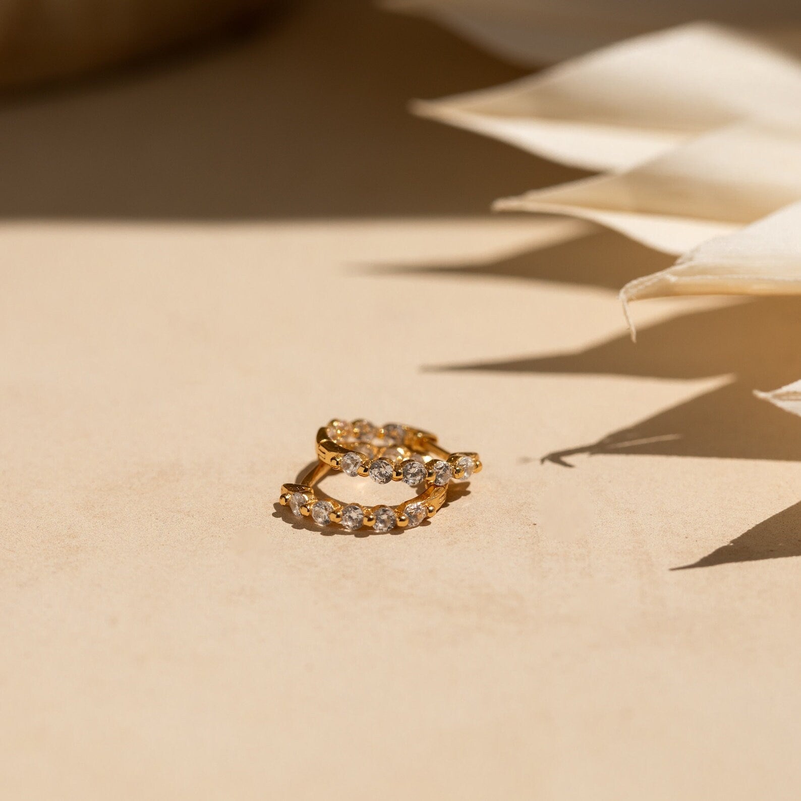 The Inside Out Huggies Set features gold hoop earrings with small clear gemstones that sparkle on a beige surface in soft, natural light, adding a subtle and refined touch to any look.