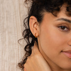 Close-up of a woman with curly hair wearing Inside Out Garnet Huggies—small gold hoop earrings—as she gently touches her neck.