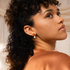 Woman with curly hair gazes upward in soft, warm lighting, wearing Inside Out Garnet Huggies and pearls.