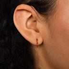 Close-up of an ear wearing the Inside Out Garnet Huggies—small gold hoop earrings with red gemstones, shown against curly dark hair—perfect for those who love delicate gemstone earrings.