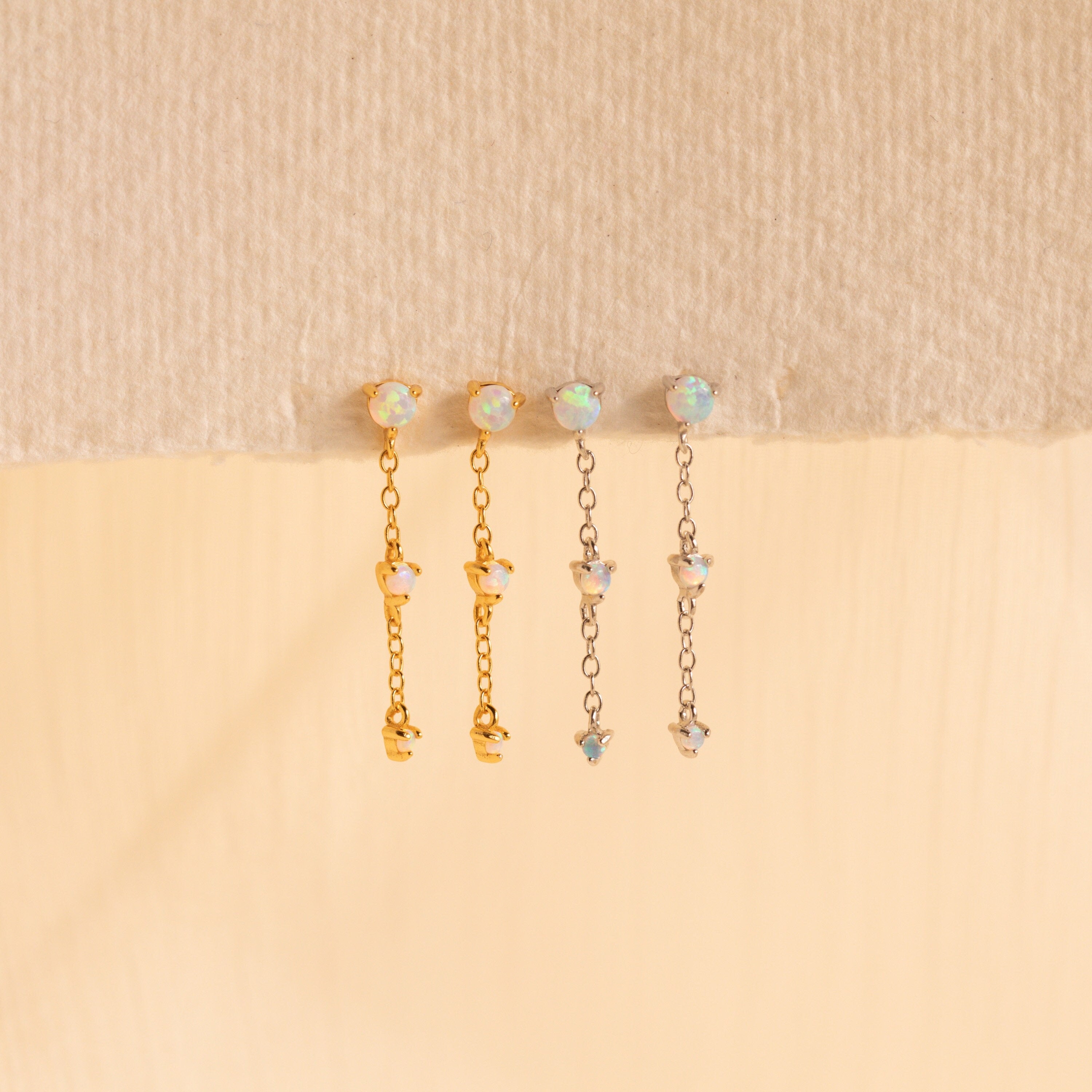 The Opal Chain Drop Earrings set includes four elegant pieces—two gold and two silver with opal-like stones—displayed on a textured surface. Perfect for bridesmaids seeking a refined, delicate touch.