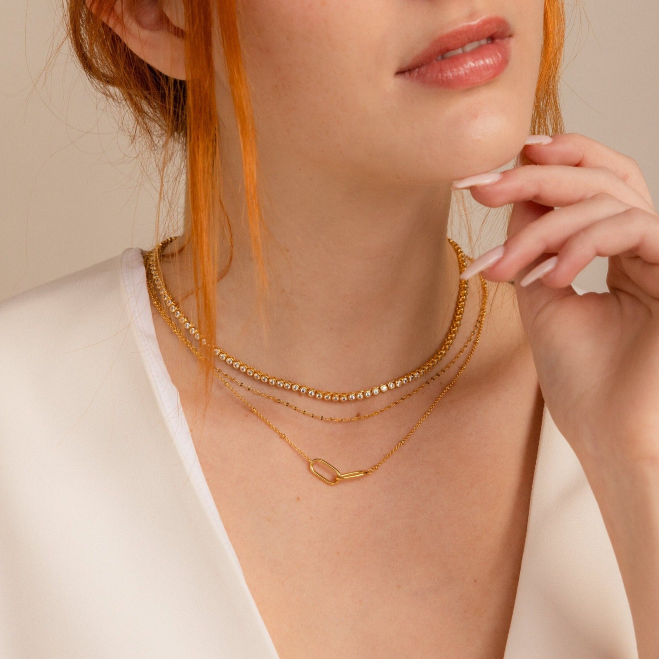 A red-haired woman in a white top wears the Diamond Bezel Tennis Necklace, a layered gold piece ideal as an elegant bridal necklace or gift for the bride, and gently touches her chin.