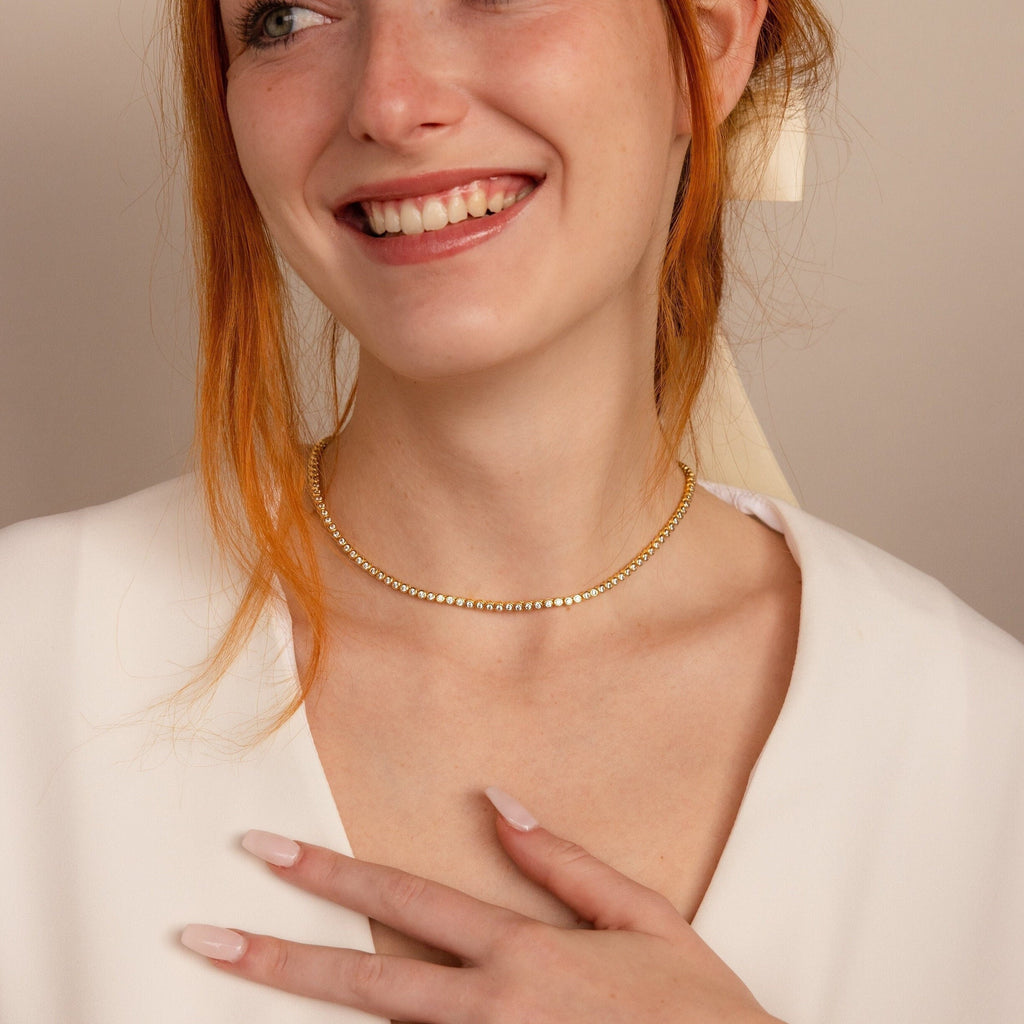 A smiling woman with red hair wears the Diamond Bezel Tennis Necklace and a white top, gently touching her chest with her manicured hand.