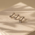 Three Diamond Flatback Studs Set in Sterling Silver, featuring clear gemstones, are displayed on textured cream fabric under soft light.