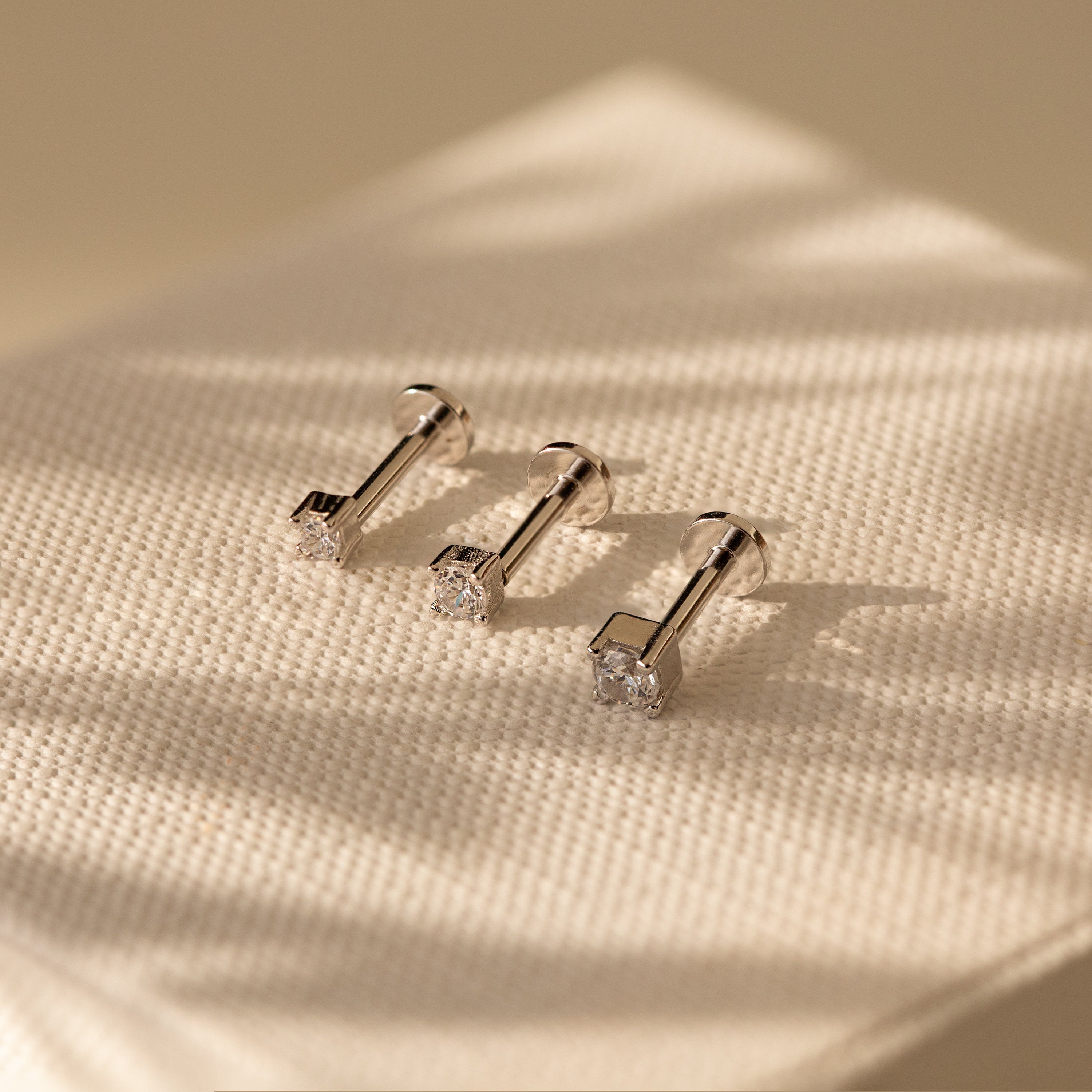 Three Diamond Flatback Studs Set in Sterling Silver, featuring clear gemstones, are displayed on textured cream fabric under soft light.