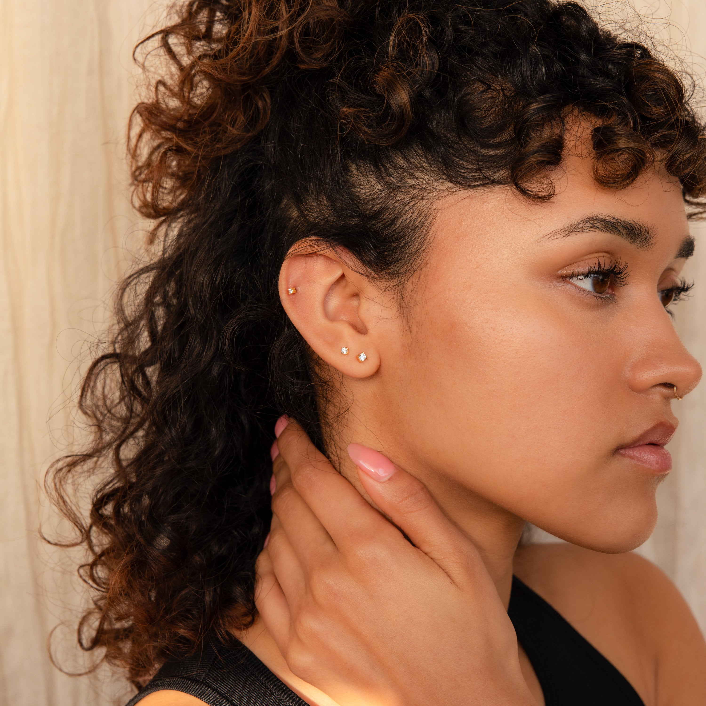A woman with curly hair and multiple ear piercings touches her neck, displaying dainty earrings and Diamond Flatback Studs Set in 18K Gold, while looking to the right against a neutral background.