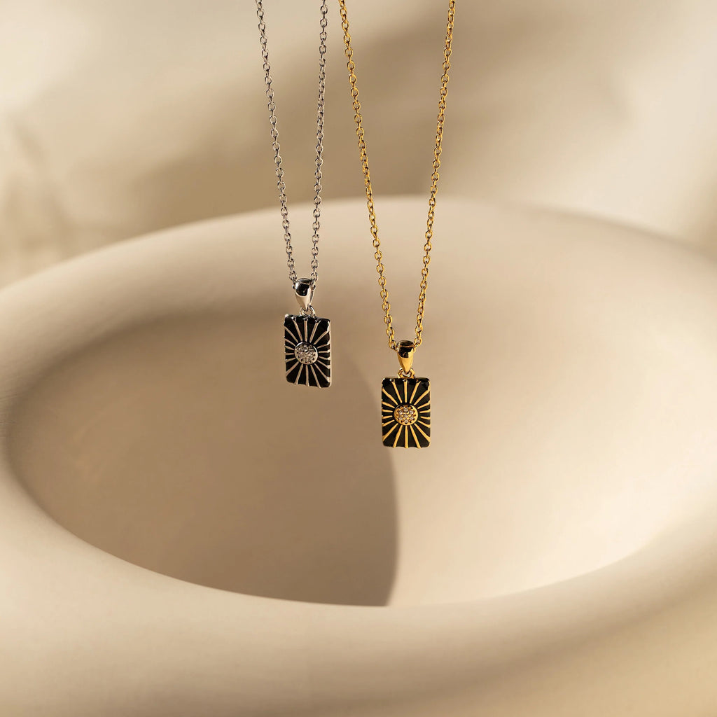 The Onyx Necklace Set features two rectangular sunburst pendants, one silver and one gold, displayed on a soft beige abstract background—ideal for minimalist jewelry layering.