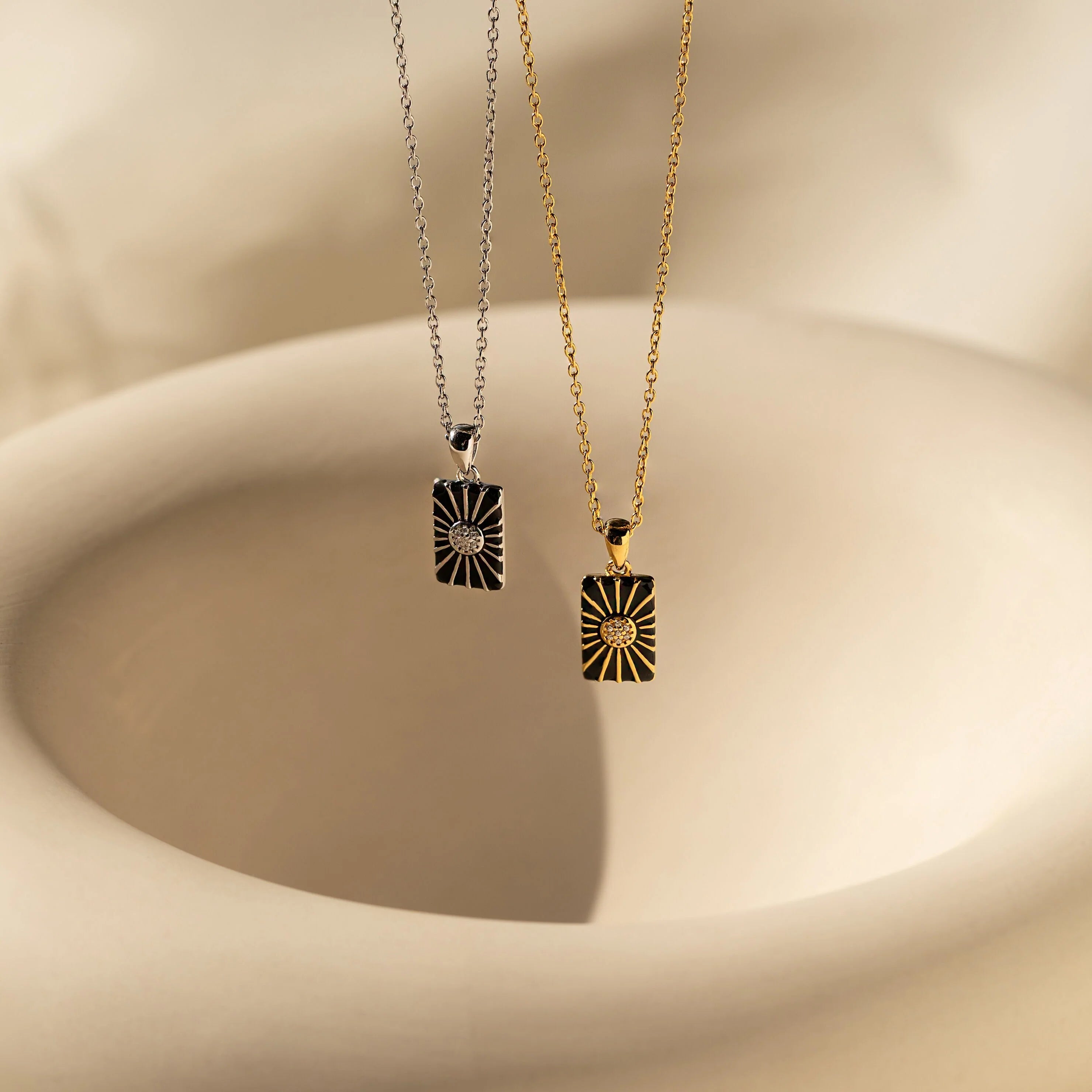 The Onyx Necklace Set features two rectangular sunburst pendants, one silver and one gold, displayed on a soft beige abstract background—ideal for minimalist jewelry layering.