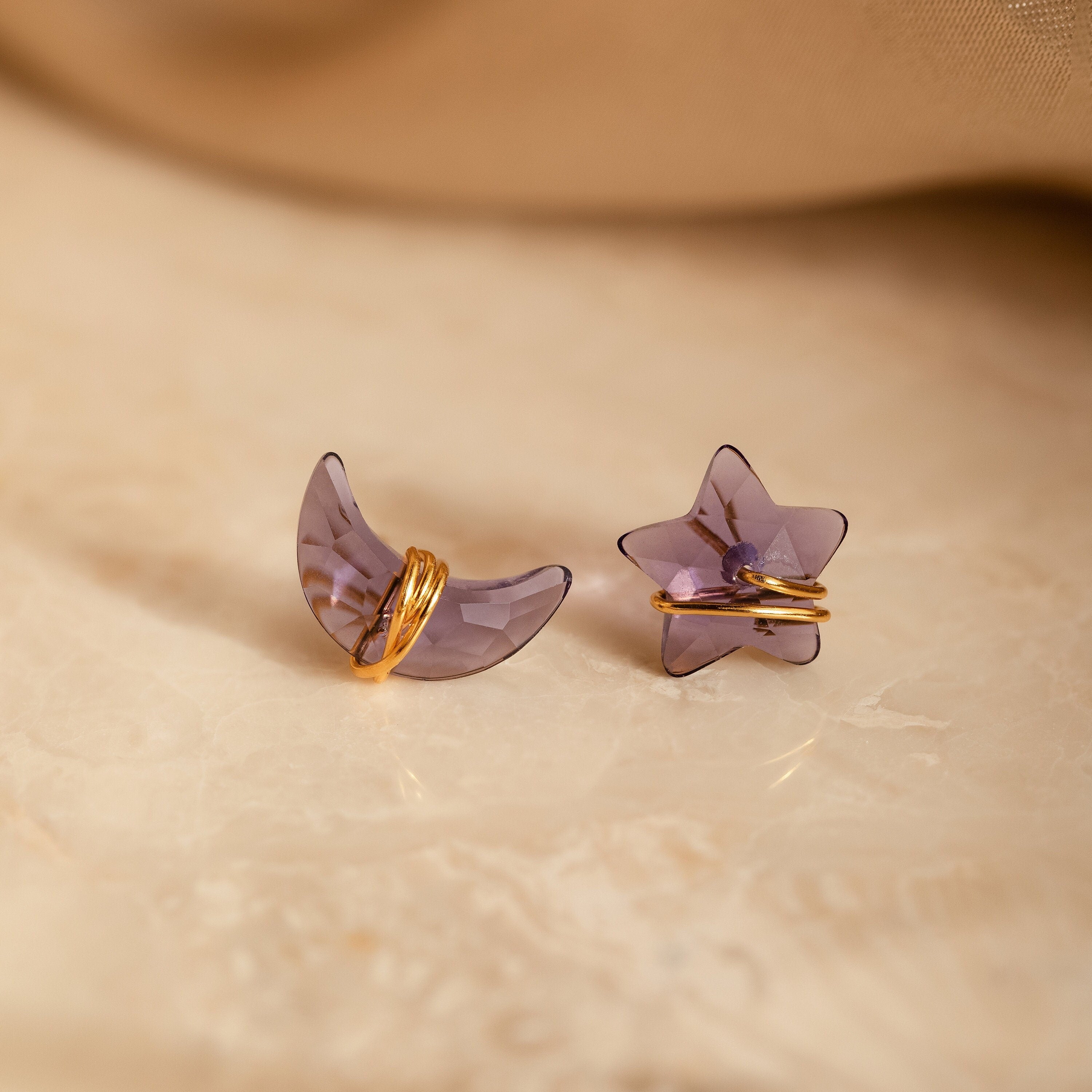 Close up of a pair of purple amethyst moon and star-shaped earrings with gold wire accents, resting on a beige marble surface.