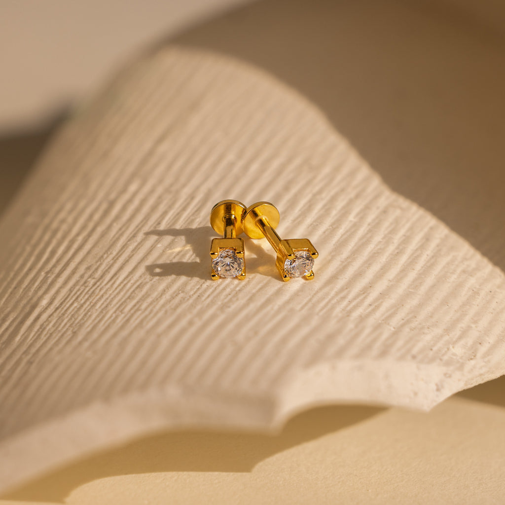 The Diamond Flatback Studs Set features gold dainty earrings with square-cut clear stones, perfect as diamond flatback studs and displayed on a textured off-white surface.