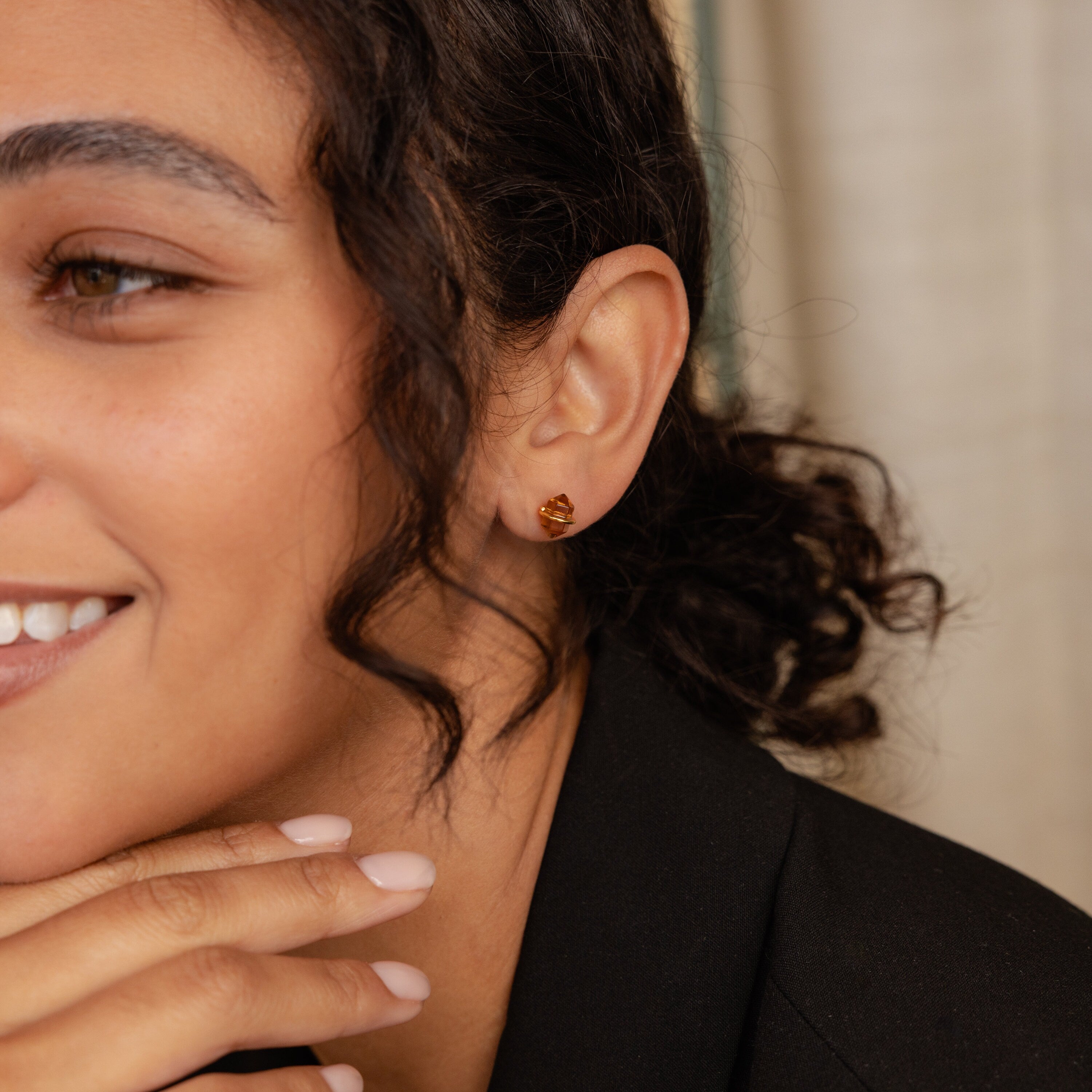 Smiling woman with curly hair, wearing Herkimer Smoky Quartz Studs and a black blazer, rests her chin on her hand.