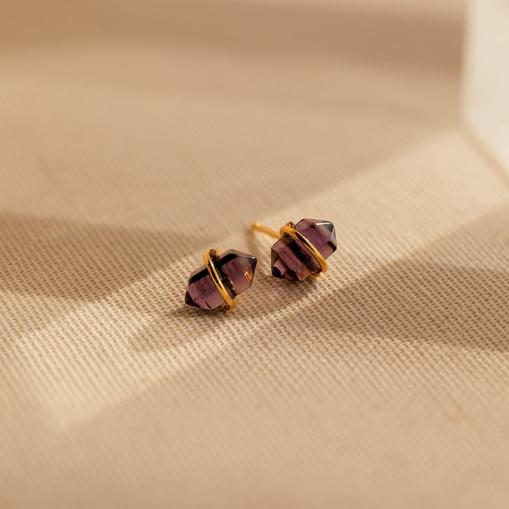 The Amethyst Herkimer Studs, featuring gold-wrapped amethyst quartz glass, rest on a beige textured surface—stunning earrings perfect for any occasion.
