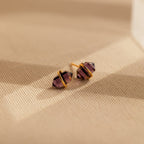 The Amethyst Herkimer Studs, featuring gold-wrapped amethyst quartz glass, rest on a beige textured surface—stunning earrings perfect for any occasion.