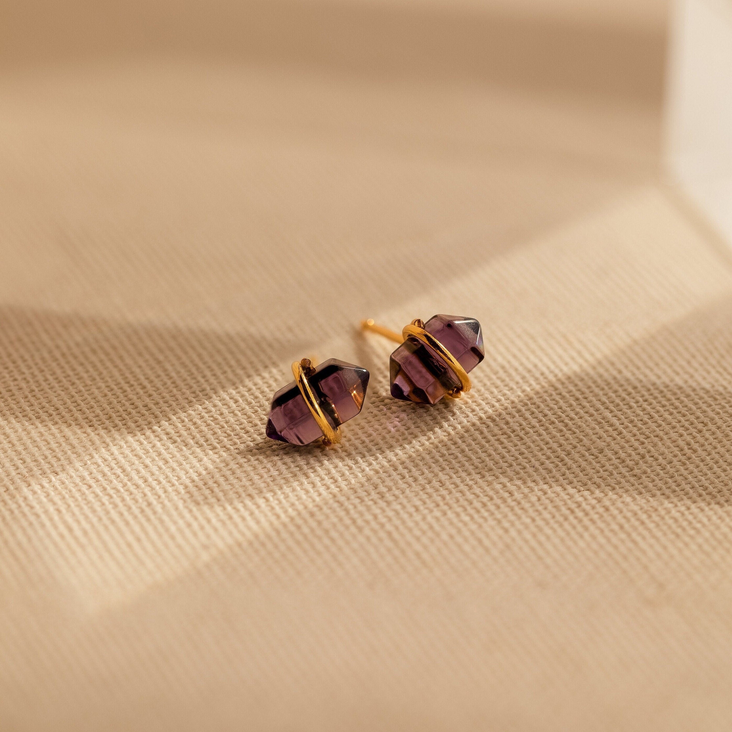 The Amethyst Herkimer Studs, featuring gold-wrapped amethyst quartz glass, rest on a beige textured surface—stunning earrings perfect for any occasion.