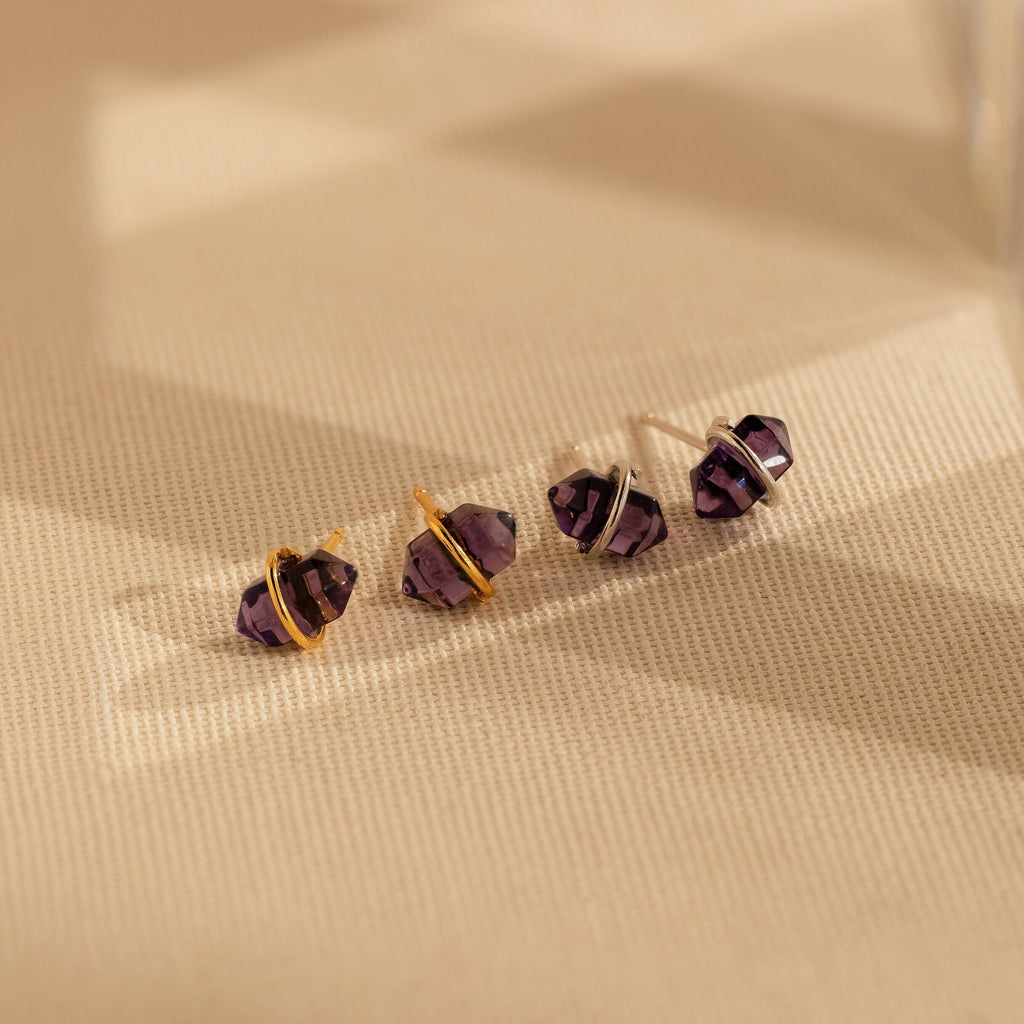 Four Amethyst Herkimer Studs, featuring gold and silver bands, are elegantly displayed on a textured beige surface.