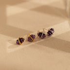 Four Amethyst Herkimer Studs, featuring gold and silver bands, are elegantly displayed on a textured beige surface.